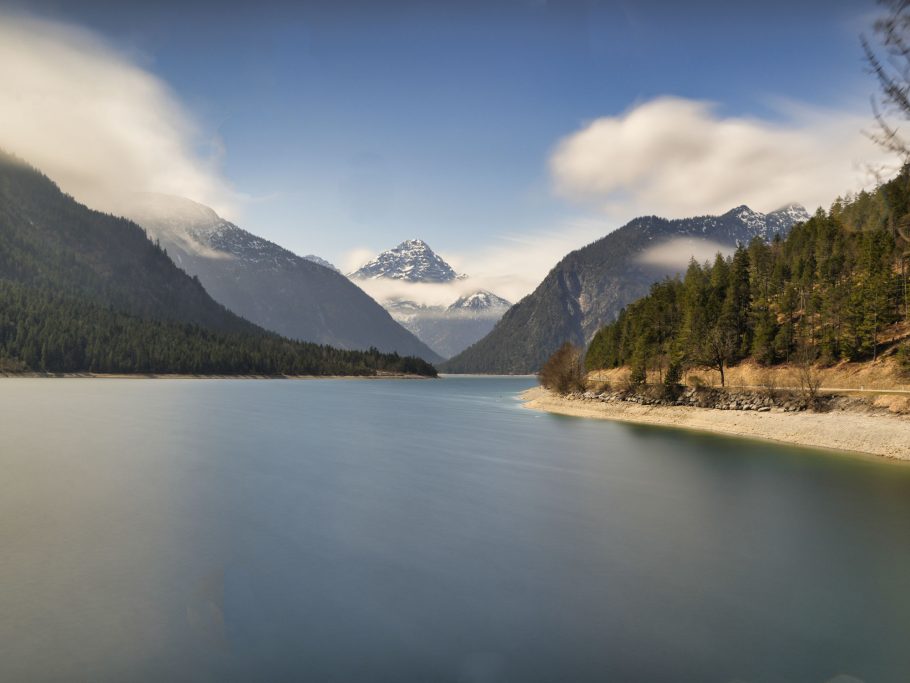 Berglandschaft mit See, umgeben von hohen Bergen und Nebel in blauer Himmel.